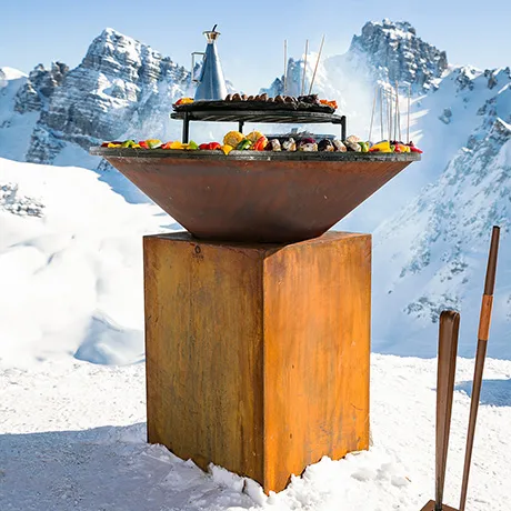 Große Feuerschale aus Cortenstahl mit buntem Grillgut vor einer Bergkulisse im Schnee