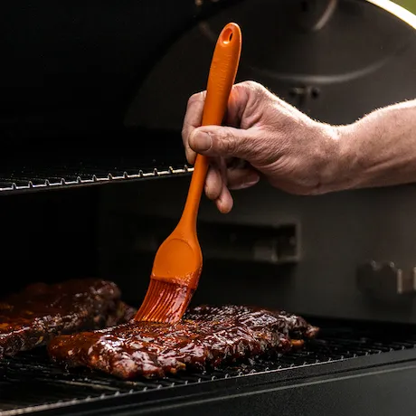 Person bestreicht Steak mit dem Silikonpinsel von Traeger in Orange auf einem Grill.