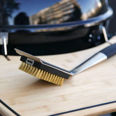 Eine Grillbürste mit einem Metallgriff und Messingborsten liegt auf einem Holzbrett neben einem Grill. Der Hintergrund zeigt unscharf einen schwarzen Grill.