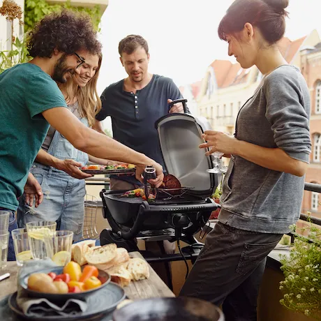 Eine Gruppe von vier Personen bei einem Grillfest auf einem Balkon, die zusammen Essen zubereiten und genießen.